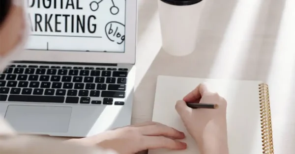 אדם כותב במחברת, לצידו מחשב נייד פתוח ועל המסך כתוב "שיווק דיגיטלי"